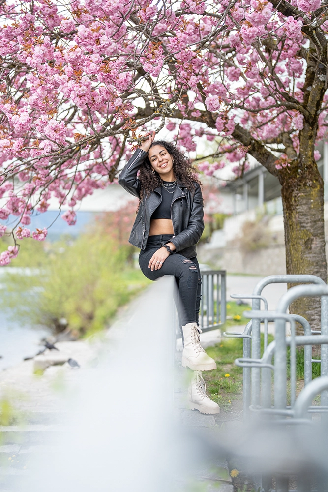 Lifestyle-Modefoto einer jungen Frau in schwarzem Outfit und Lederjacke, sitzend unter blühendem Kirschblütenbaum am Ufer – Fashion & Portraitfotografie Villach, Kärnten