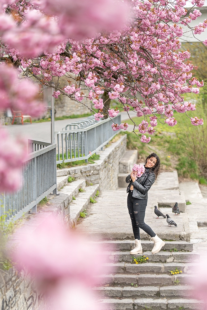 Junge Frau in schwarzer Jeans, Lederjacke und Boots posiert mit Kirschblütenstrauß auf Steinstufen – Frühlings-Mode- und Portraitshooting in Villach, Kärnten