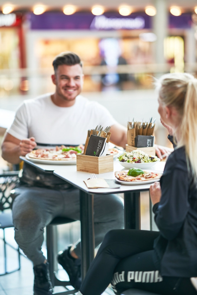 Zwei Gäste im Restaurant Al Dente im Atrio Villach im Atrio Villach essen Pizza und Salat an einem Tisch; Besteckspender im Vordergrund.