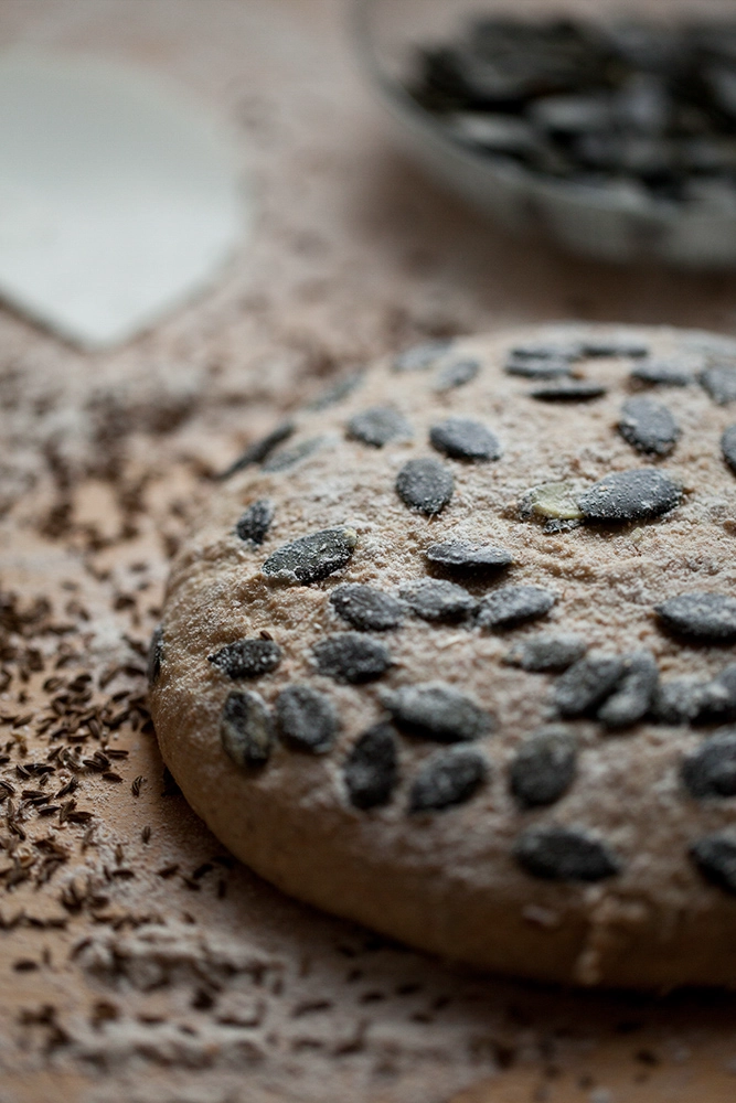 Runder Brotteig mit Kürbiskernen bestreut; Kümmel auf der Arbeitsfläche.