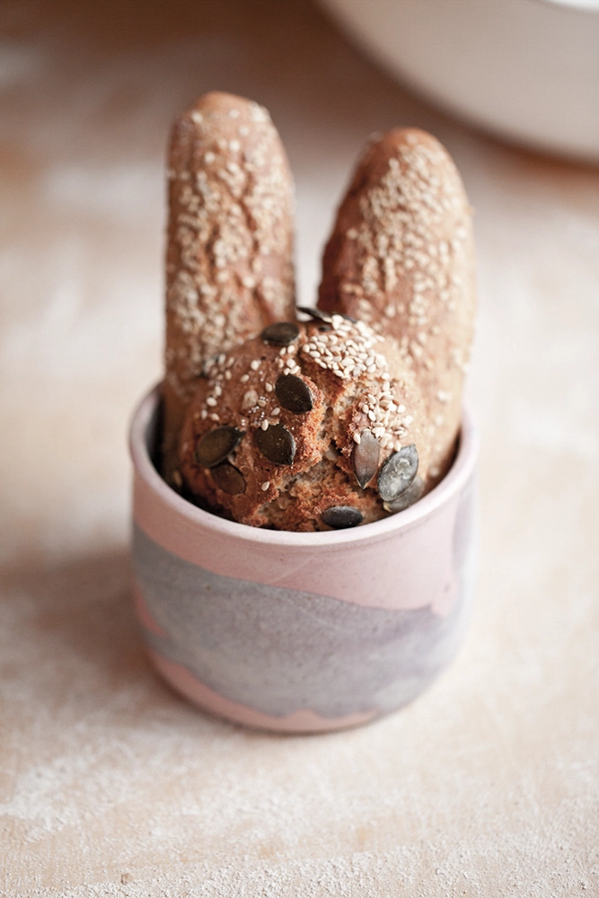 Kleines Gebäck mit Sesam und Kürbiskernen in einem rosafarbenen Becher; stehende Teigstücke.