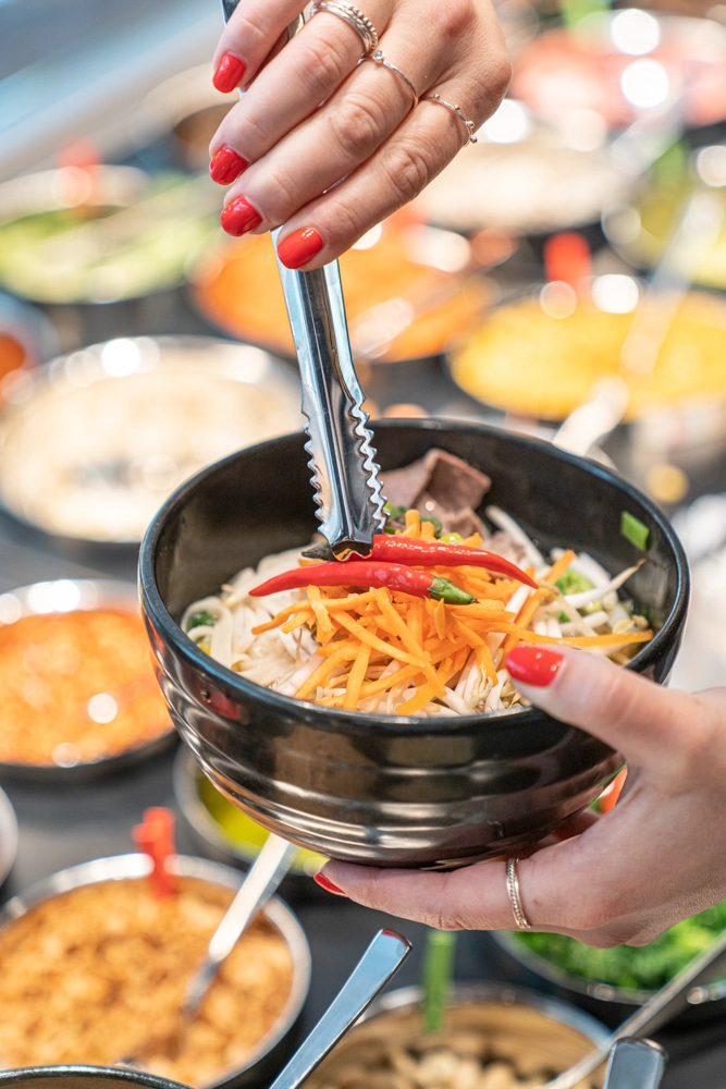 Nahaufnahme einer asiatischen Bowl mit Nudeln, Karotten und Chili im Restaurant My Indigo im Atrio Villach; eine Zange gibt Chilli in die Bowl, diverse frische Zutaten im Hintergrund.