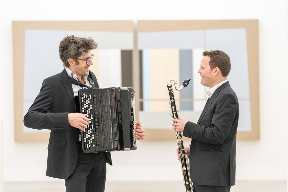 Werbefoto Duo-Klak – Akkordeonspieler und Bassklarinettist im MMKK Klagenfurt, im Gespräch vor abstrakter Kunst.