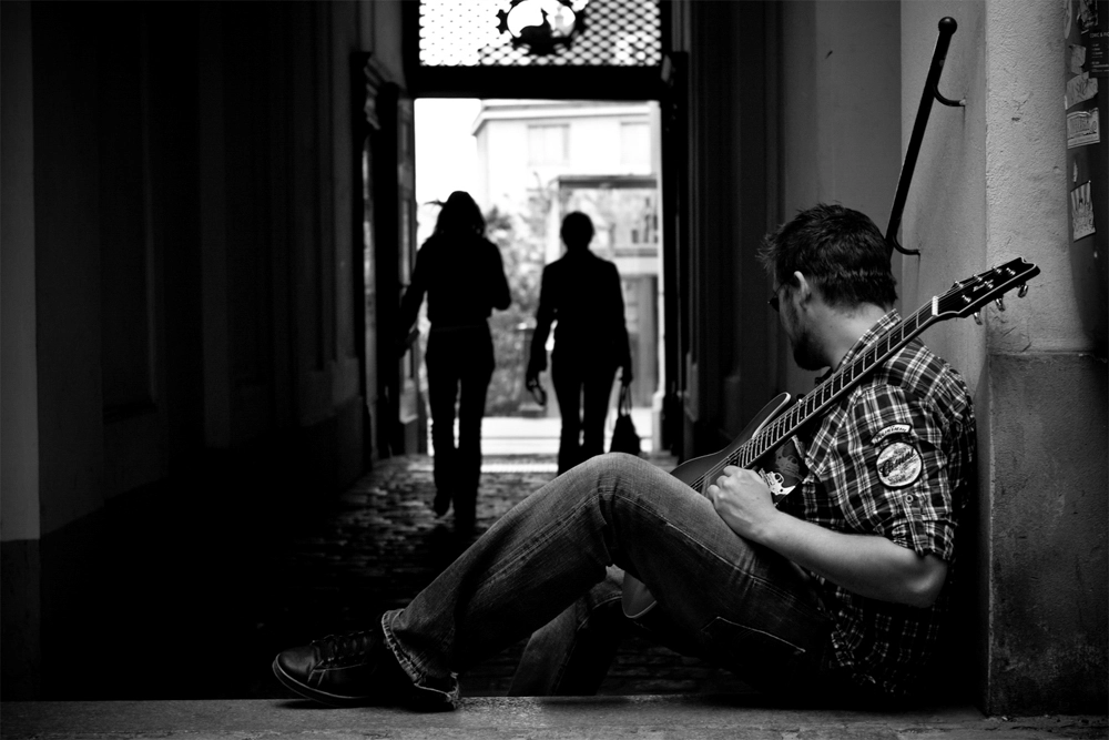 Werbefoto Schwarz‑Weiß‑Foto: Gitarrist im Durchgang, Silhouetten von Passant:innen im Hintergrund in der Stadt Wien.