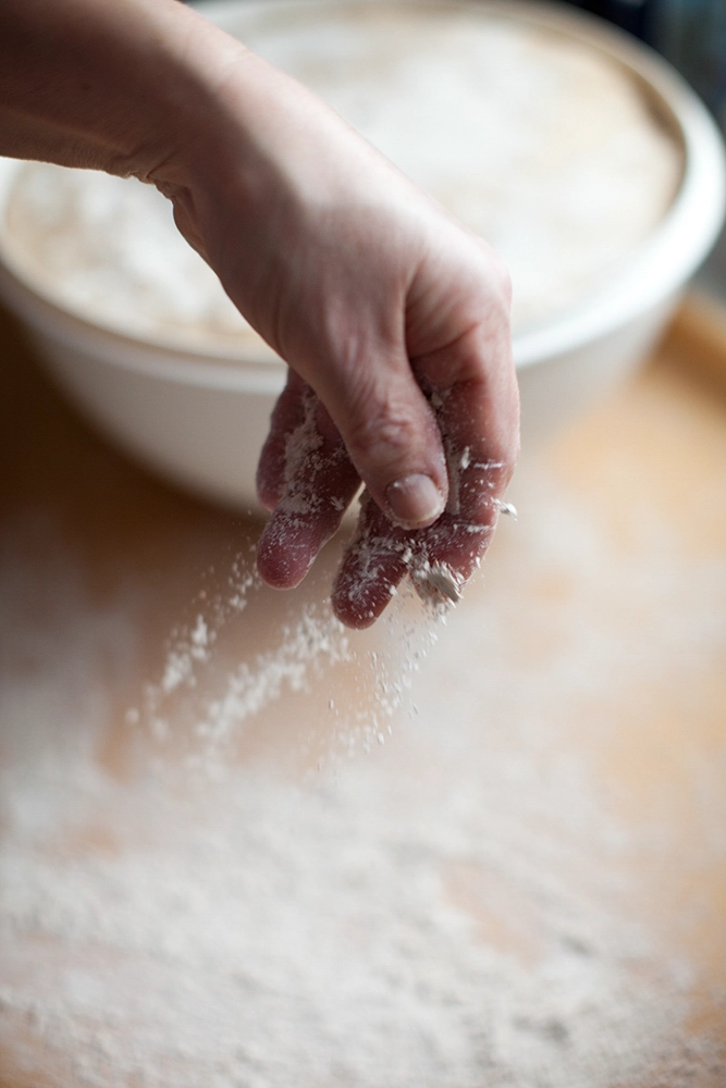 Hand streut Mehl auf die Arbeitsfläche; Schüssel mit Teig unscharf im Hintergrund.